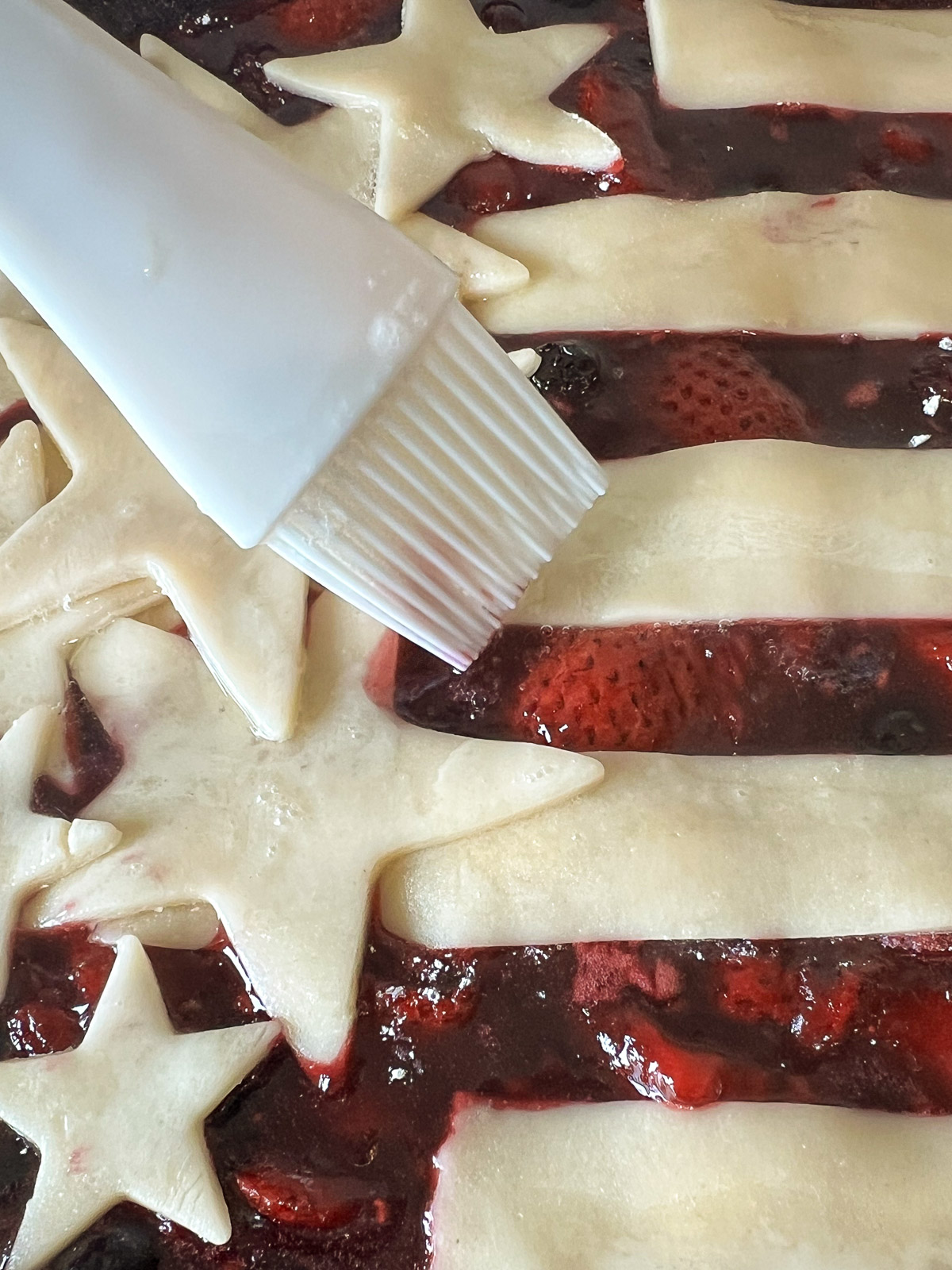 Egg wash being brushed on pie crust with sparkling sugar for berry cobbler