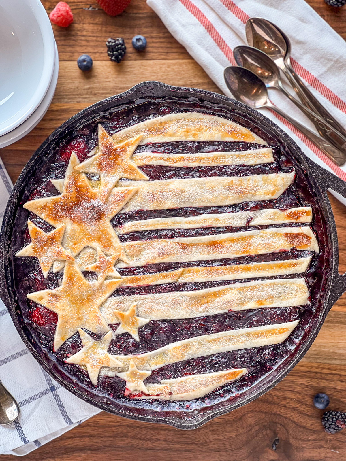 Cast iron skillet berry cobbler with golden baked pie crust stars and stripes over bubbling mixed berry filling