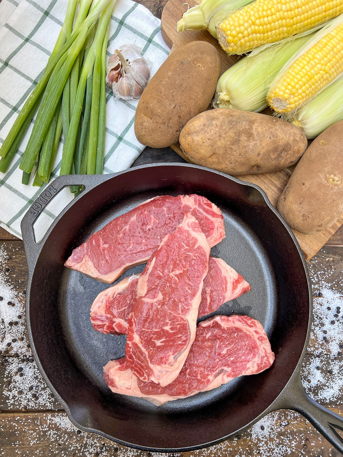 Raw strip steak in a cast iron skillet with corn, potatoes, and green onions in the background.