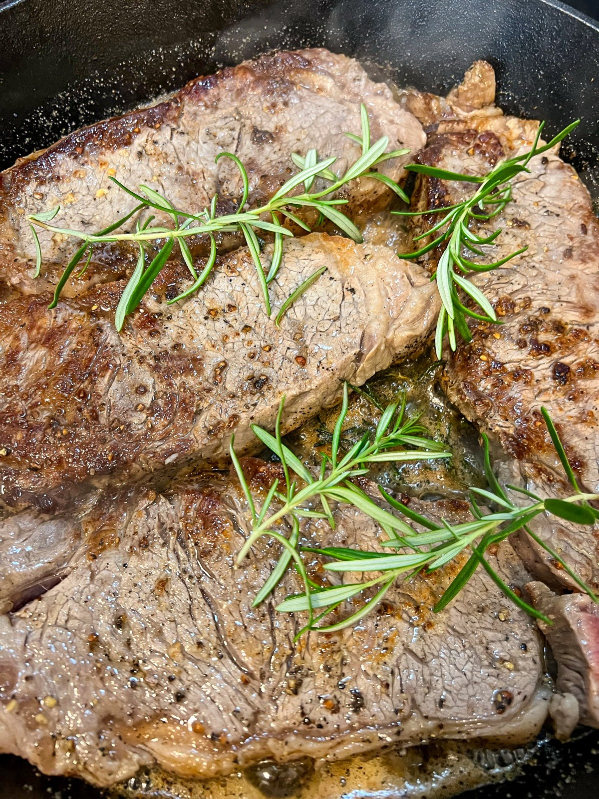 Branded image of cooked strip steaks in cast iron garnished with fresh rosemary.