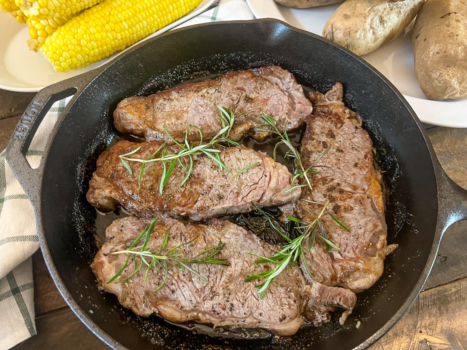 Steak, baked potato, and corn on the cob on a white plate with those items in individual vessels in the background.
