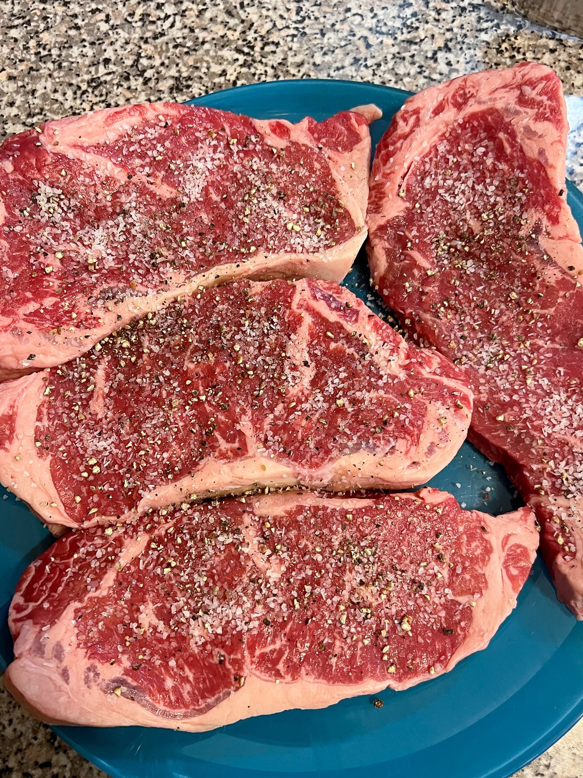 4 New York strip steaks on a plate coming to room temperature seasoned with salt and pepper.
