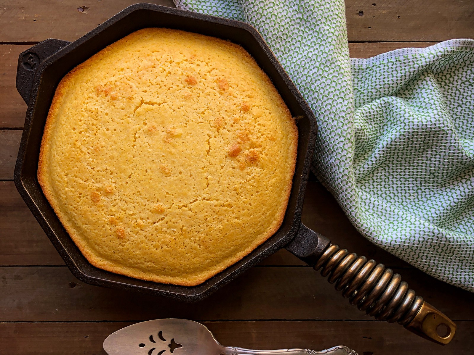 Cast Iron Sweet Cornbread freshly baked in a skillet with a towel and slice server.