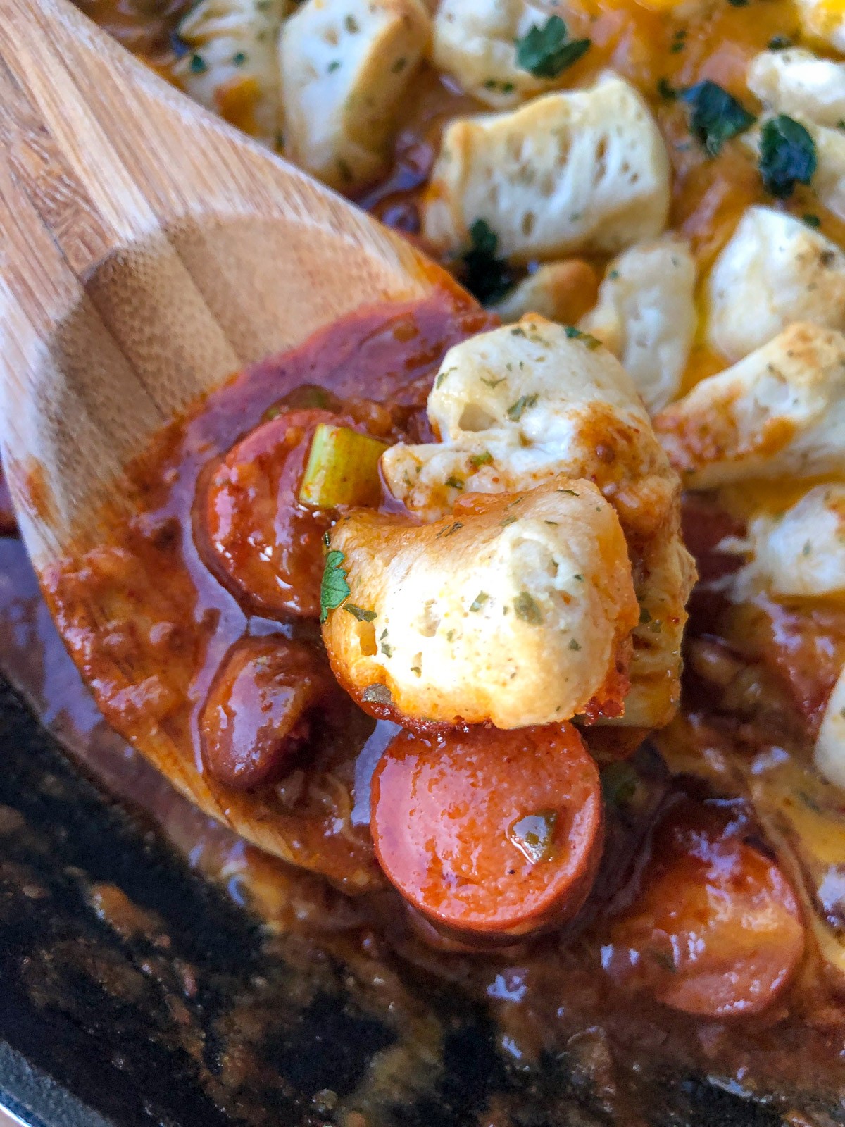 A scoop of hot dogs, chili, and garlic butter biscuits on a wooden spoon.