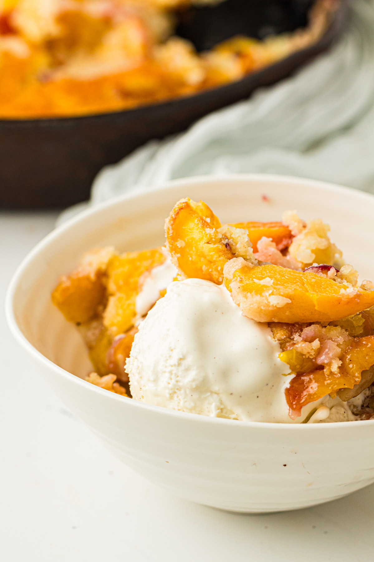 Dutch Oven Peach Cobbler in a white bowl with ice cream sitting next to a cast iron skillet. 