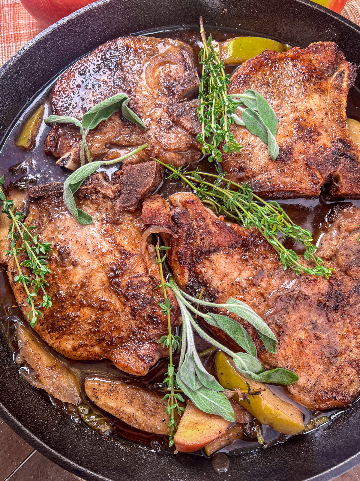 Cast iron skillet with four pork chops atop cooked apples and onions with sprigs of fresh herbs as a garnish.