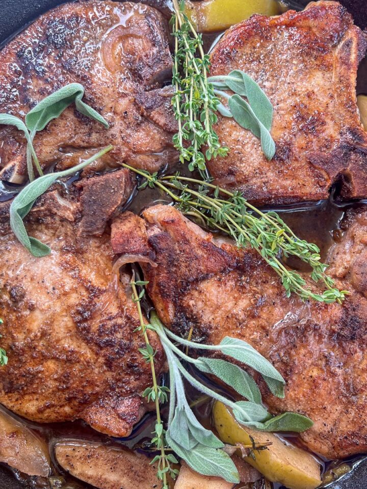 Pork chops cooking in a cast iron skillet with apples and fresh herbs.