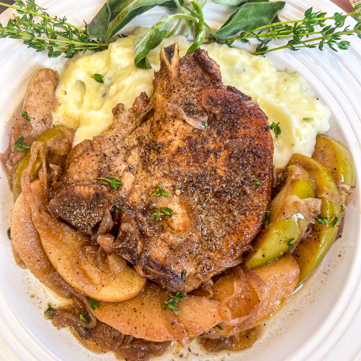 A plated pork chop served over mashed potatoes with apples, onions, and fresh herbs.