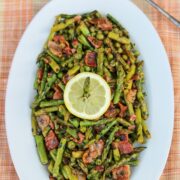 Asparagus and peas with bacon and herbs on a white oval platter garnished with a lemon slice.