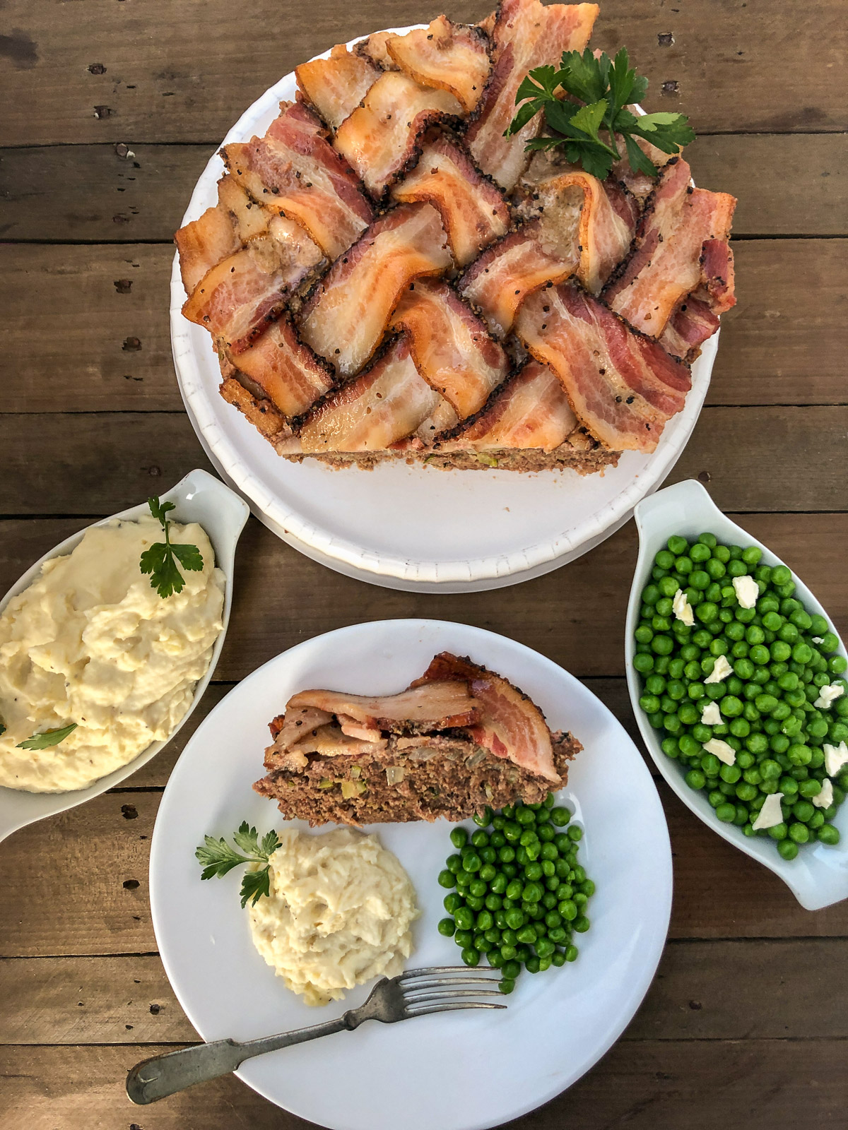 Full spread of bacon wrapped meatloaf with a plated slice, mashed potatoes and peas