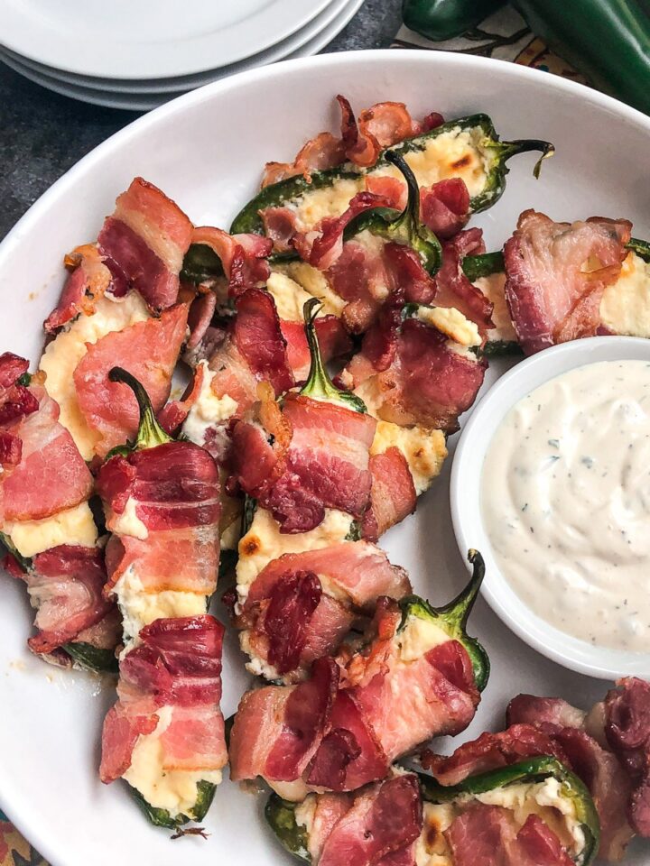 Bacon wrapped jalapeno poppers on a white plate with ranch dip