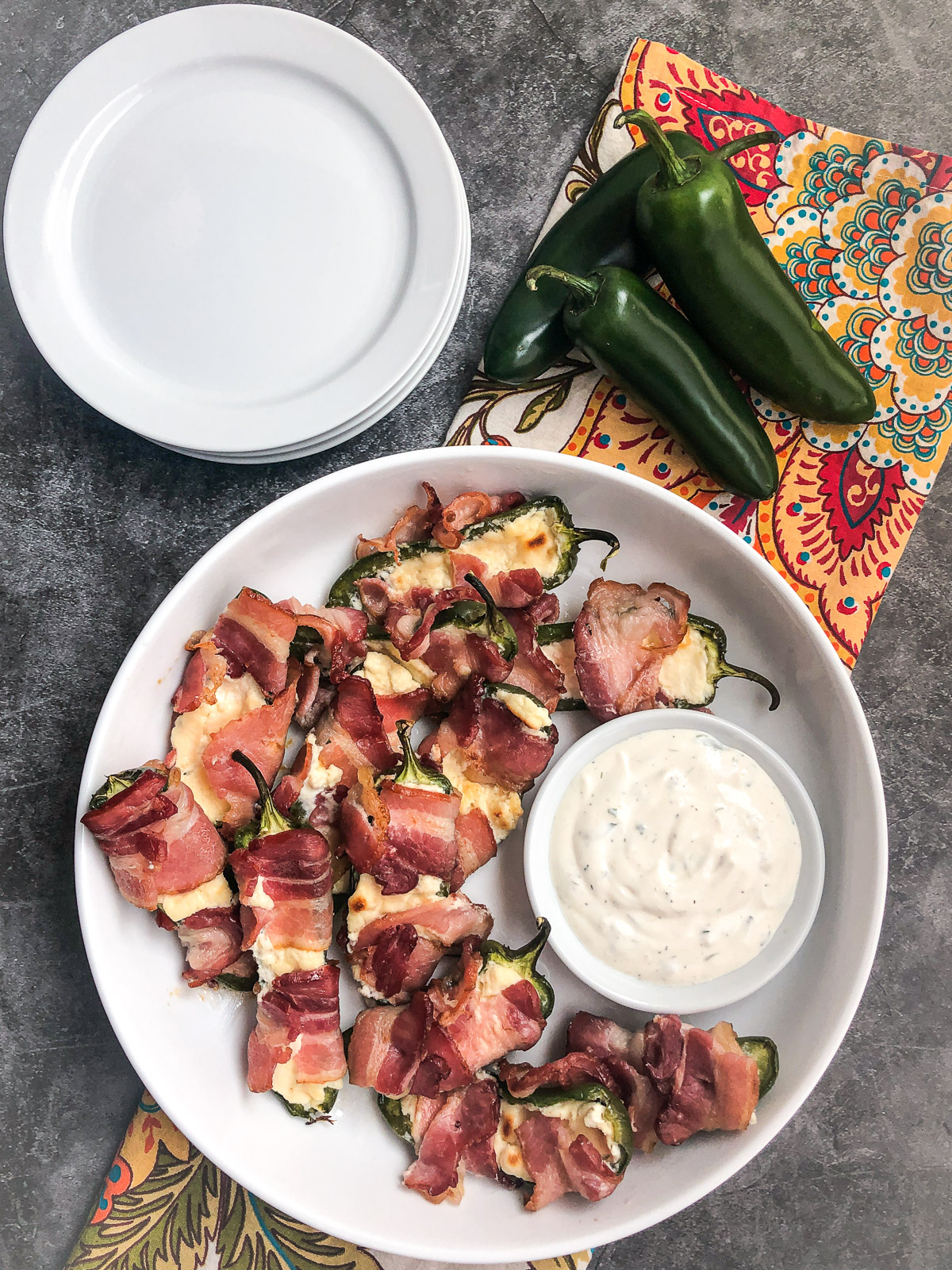 A white plate of bacon wrapped jalapeno poppers with ranch dip, fresh jalapenos and plates in the background