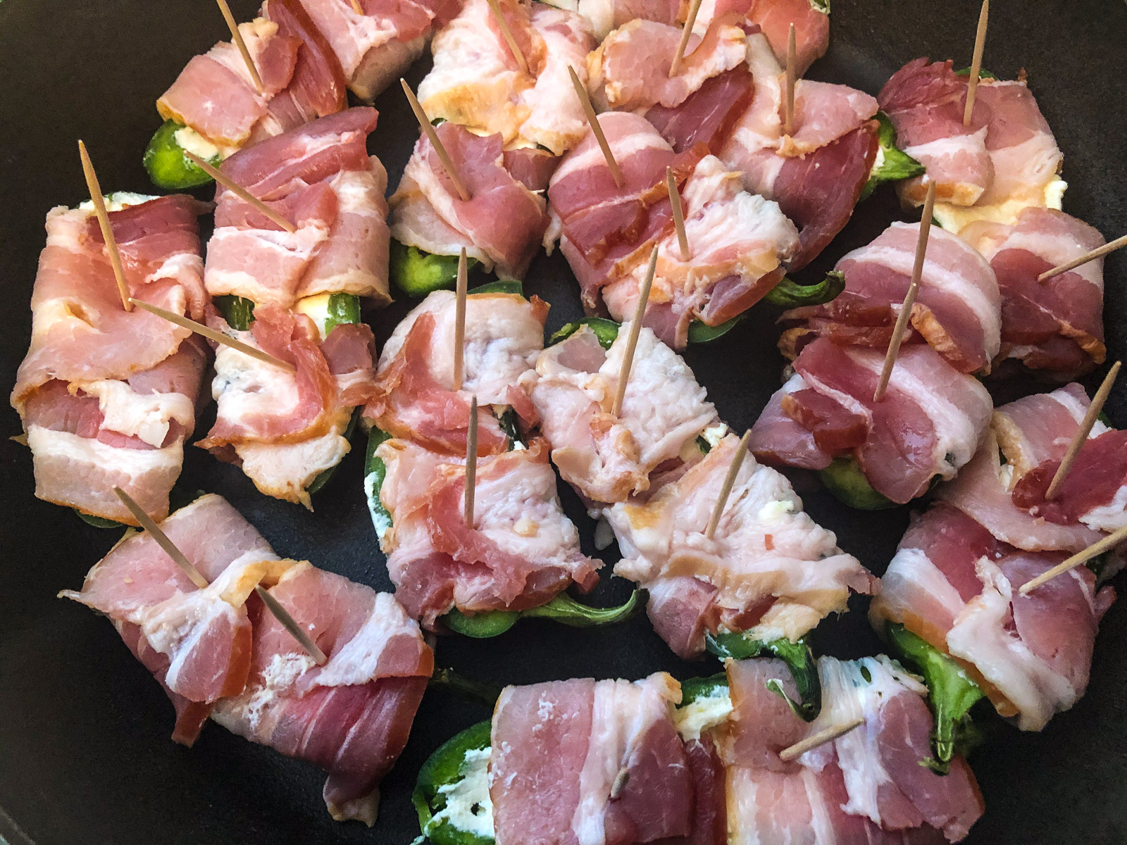 Bacon wrapped jalapeno poppers arranged in a cast iron skillet with toothpicks before baking