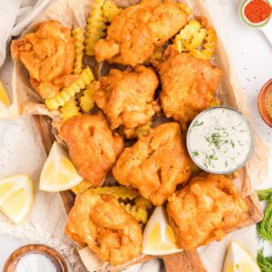 Beer battered fish on a wooden platter with fries, tartar sauce, and lemon wedges