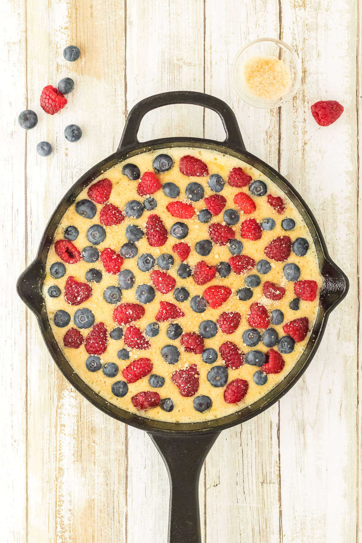 Fresh raspberries and blueberries pressed into brown sugar cake batter in a cast iron skillet with turbinado sugar