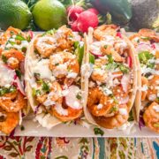 A platter with four chili lime shrimp tacos fully loaded with cabbage, radishes, cilantro, jalapenos, queso fresco, and creama.