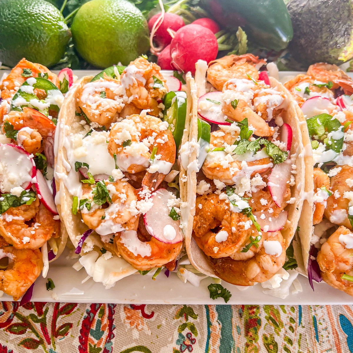 A platter with four chili lime shrimp tacos fully loaded with cabbage, radishes, cilantro, jalapenos, queso fresco, and creama.
