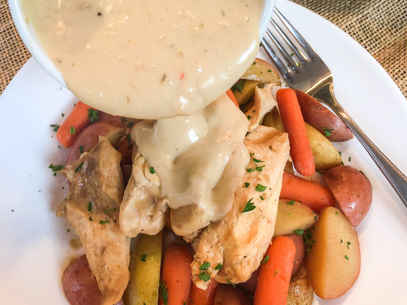 Creamy gravy being poured over plated Italian dressing chicken with carrots and potatoes.