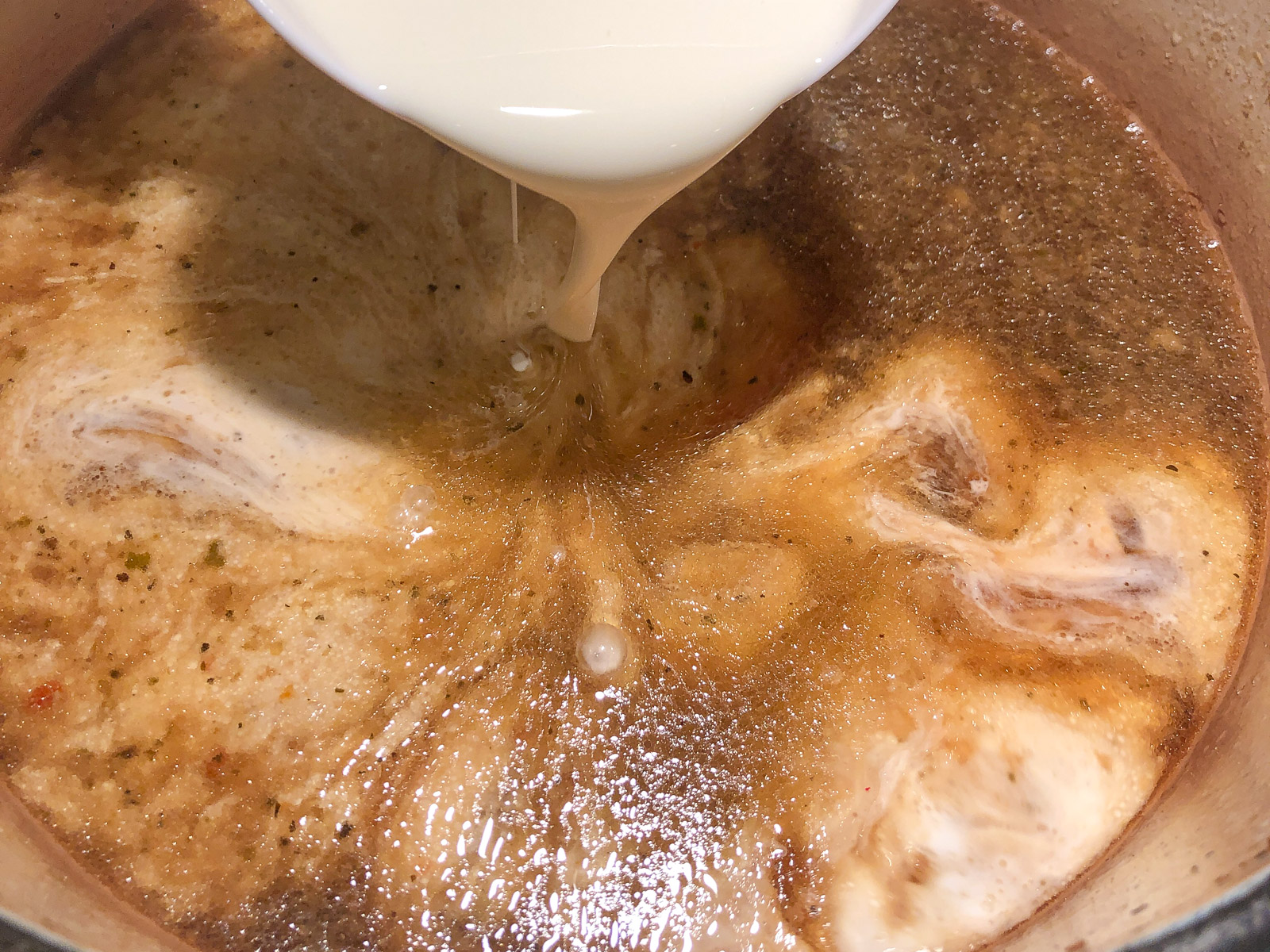 Heavy cream being poured into the remaining cooking liquid in the Dutch oven to make gravy.