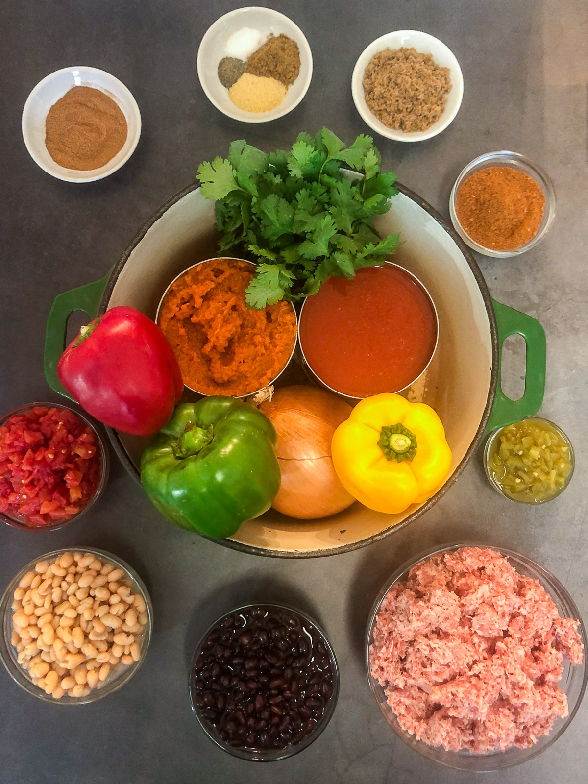 All the ingredients for Dutch oven pumpkin chili arranged in and around a Dutch oven including ground sausage, bell peppers, pumpkin puree, tomato sauce, beans, and spices.