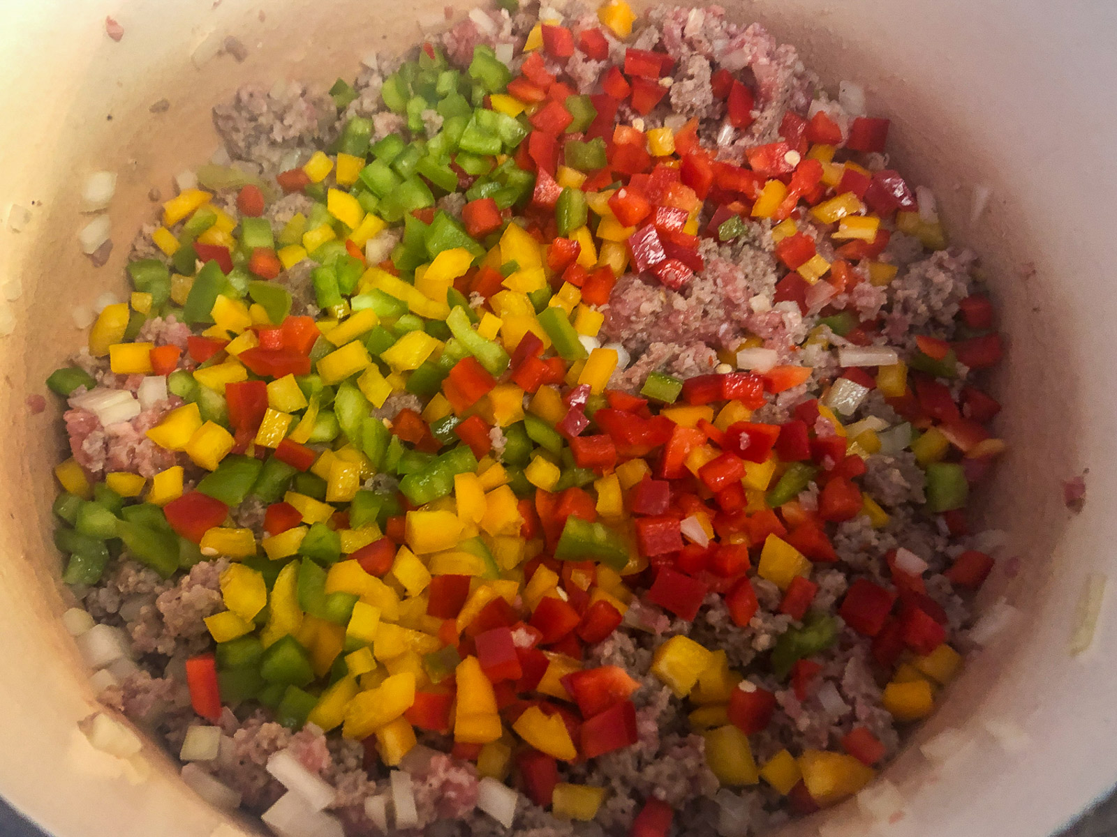 Diced red, yellow, and green bell peppers added on top of browned sausage and onions in a Dutch oven.