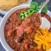 A spoonful of pumpkin chili lifted from a white bowl showing thick, hearty texture with beans and ground sausage.