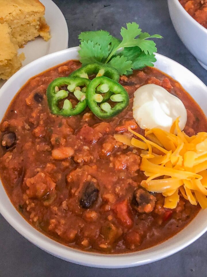 Pumpkin chili served in a white bowl garnished with jalapeños, cilantro, sour cream, and shredded cheddar cheese with cornbread on the side.