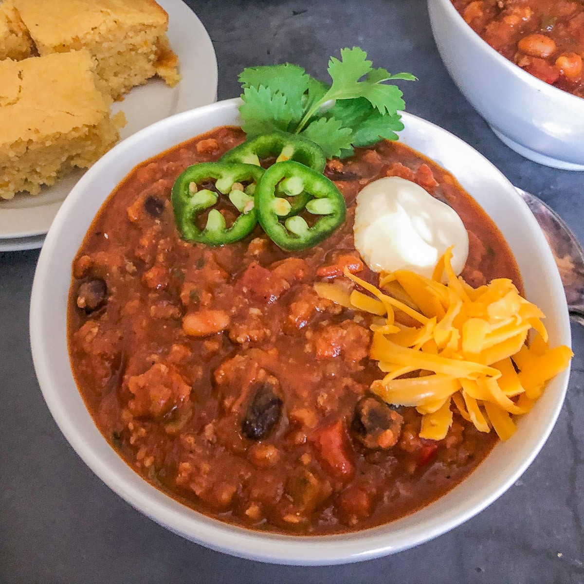 Pumpkin chili served in a white bowl garnished with jalapeños, cilantro, sour cream, and shredded cheddar cheese with cornbread on the side.