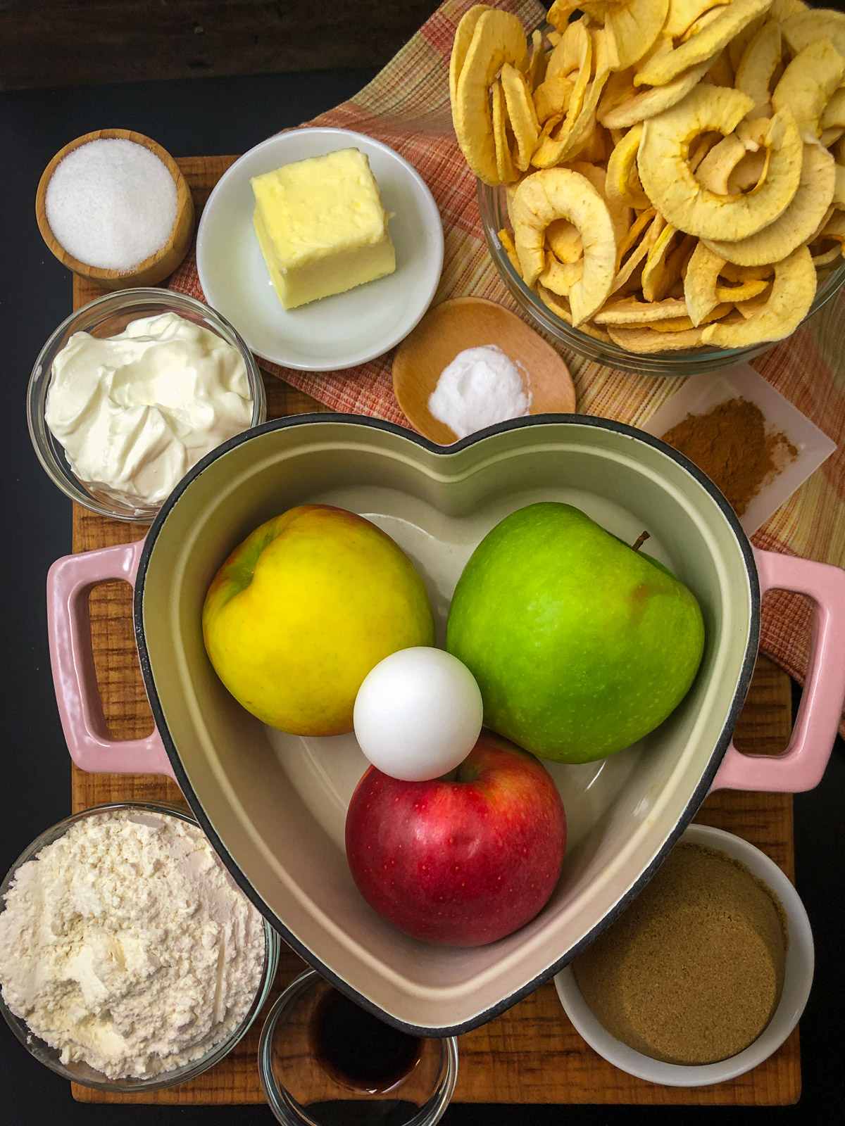 All ingredients for Dutch oven apple cake arranged around a heart-shaped cast iron Dutch oven with three fresh apples inside