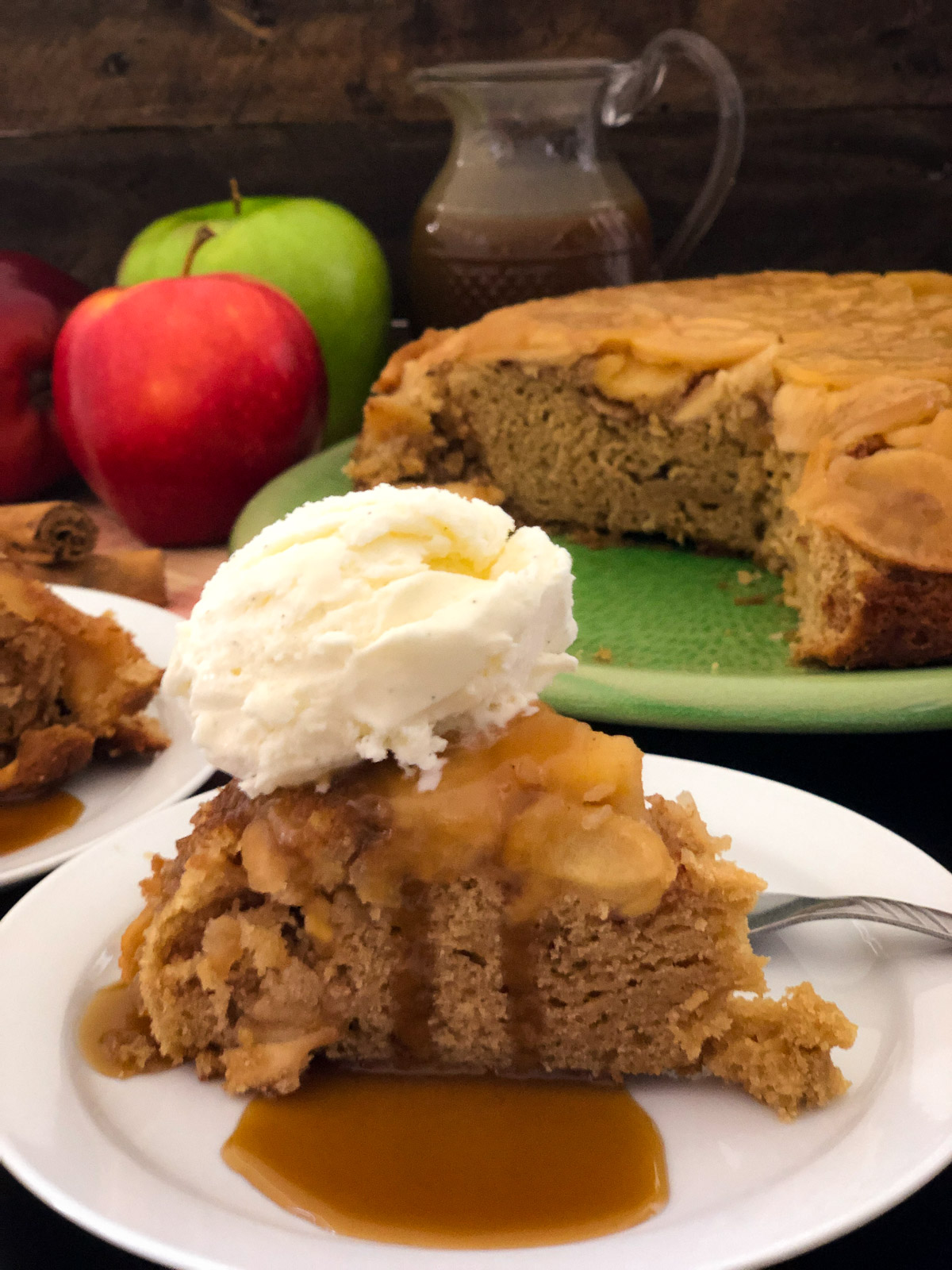 A slice of Dutch oven apple cake on a white plate with caramel sauce and vanilla ice cream, whole cake on green platter in background