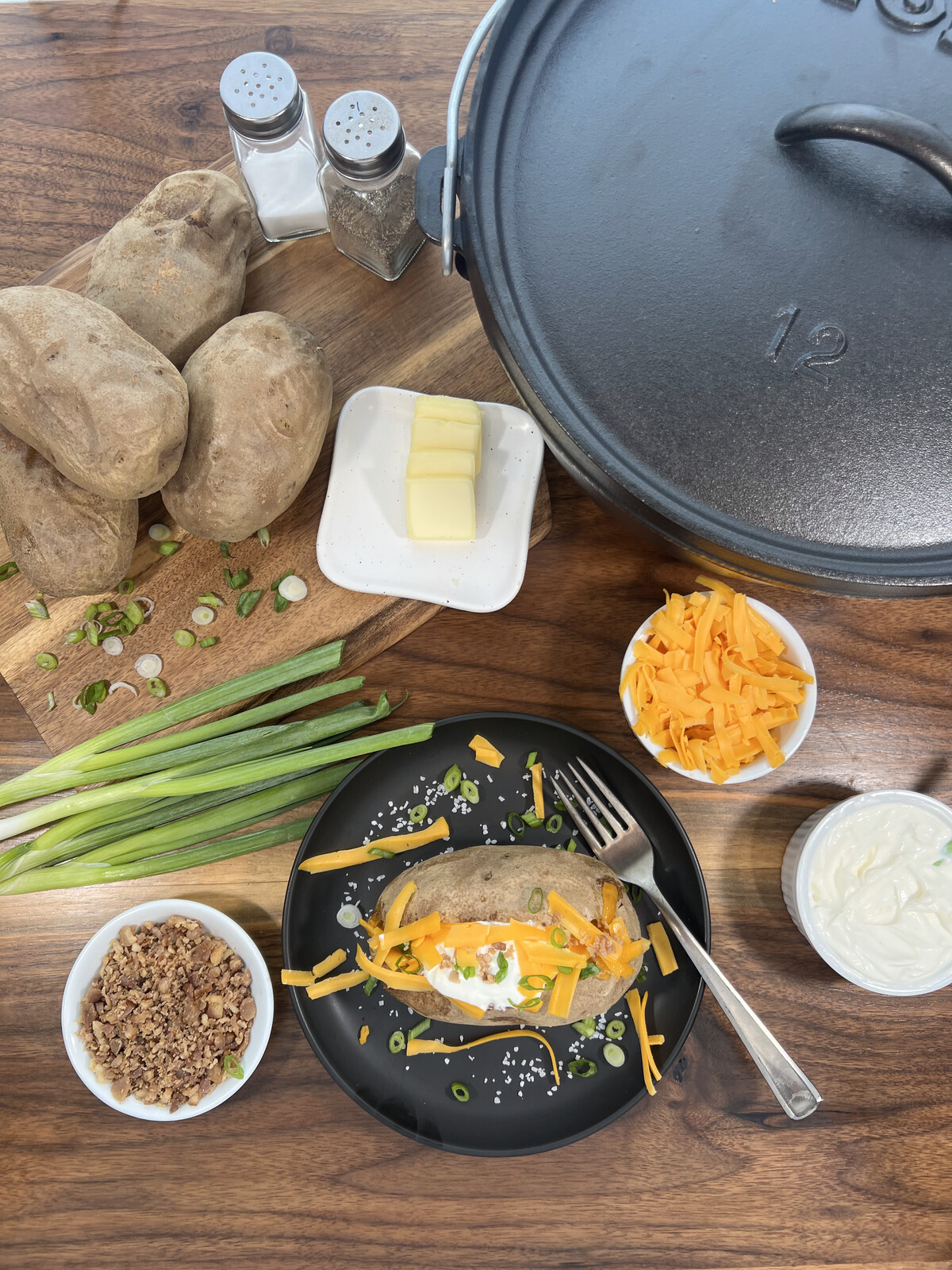 Loaded baked potato on a plate with cheese, sour cream, bacon, and green onions next to a cast iron dutch oven