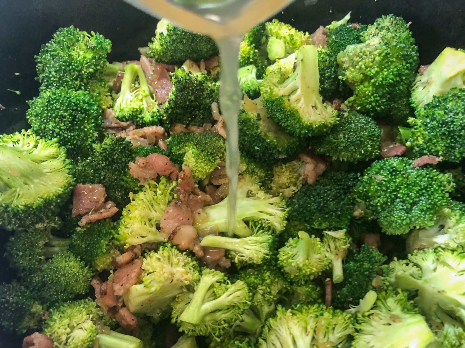 Chicken broth being poured over broccoli, bacon, and onions in the Dutch oven