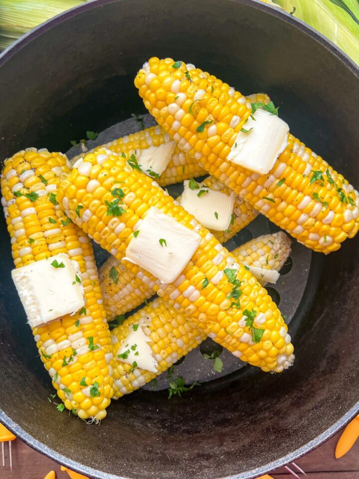 Freshly steamed corn on the cob in a dutch oven with butter and chopped parsley.
