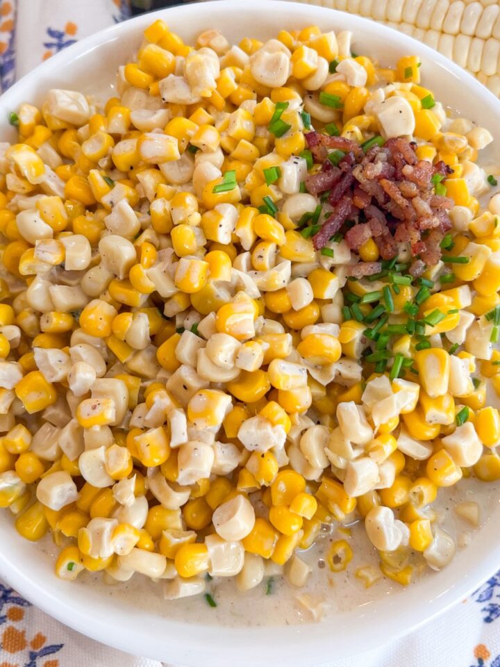 Creamy corn in a white bowl garnished with bacon crumbles and chives with spoons and fresh corn cobs in the background.