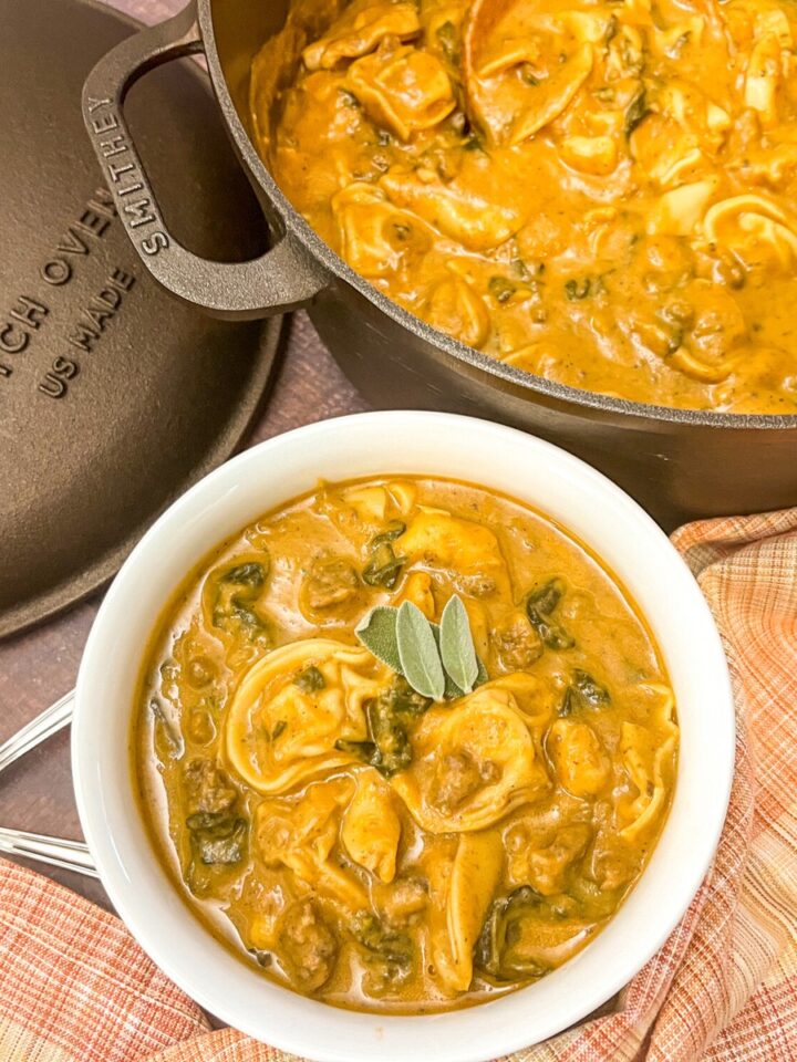 A hearty serving of butternut tortellini sausage soup in a white bowl with a rest in the dutch oven in the background.
