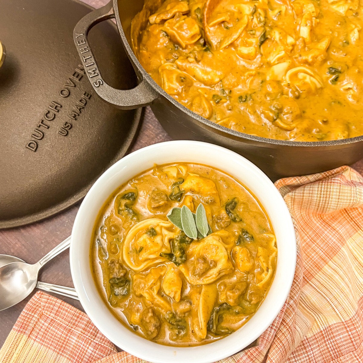 A hearty serving of butternut tortellini sausage soup in a white bowl with a rest in the dutch oven in the background.