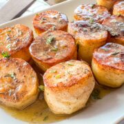 Fondant potatoes on a white serving platter with pan sauce drizzled over and fresh thyme garnish