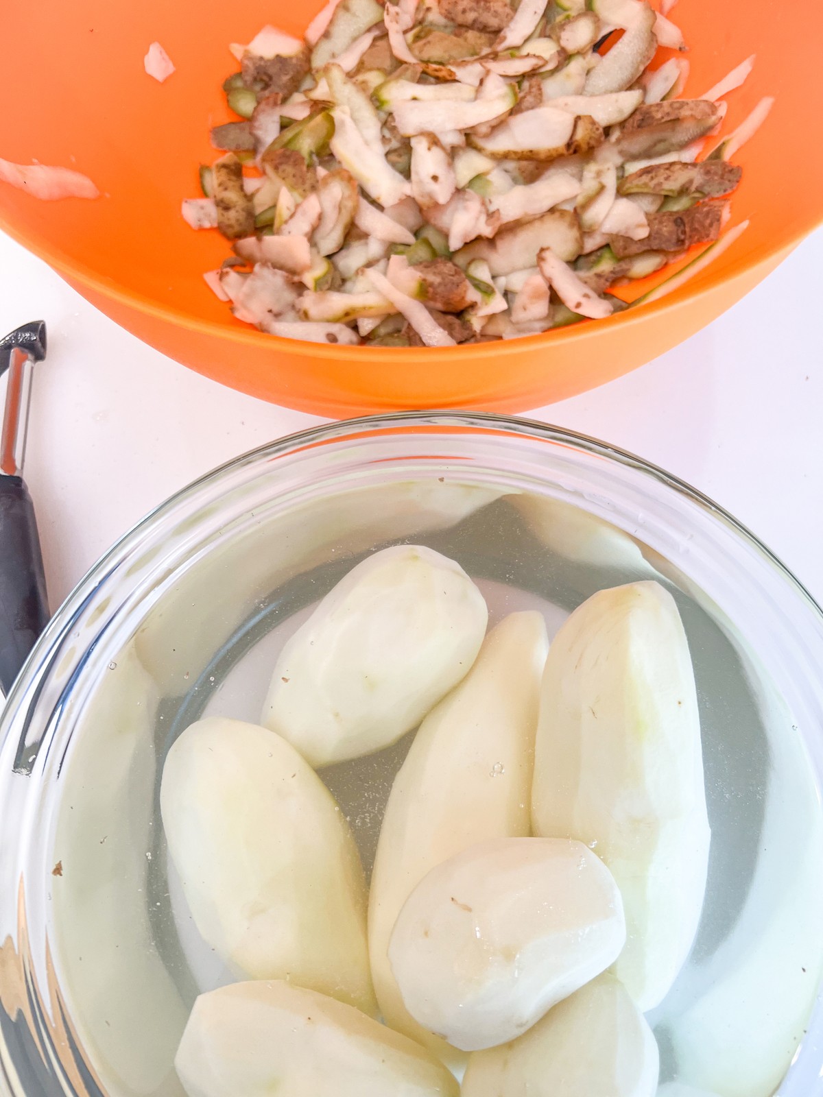 Peeled potatoes in a bowl of cold water to prevent browning and the peels in an orange bowl for composting.