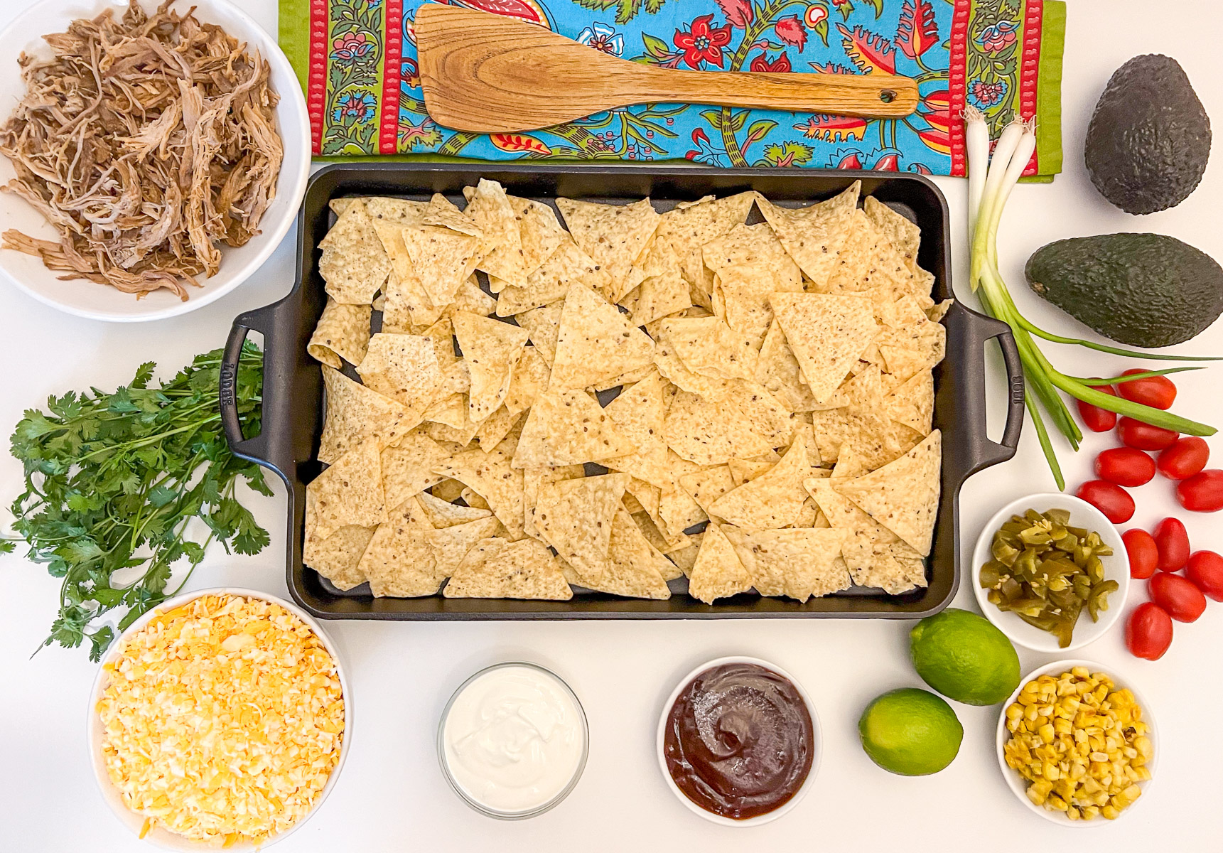 Flat lay of pulled pork nachos ingredients: pulled pork, tortilla chips on a cast iron baking sheet, shredded cheese, cilantro, tomatoes, jalape\u00f1os, roasted corn, barbecue sauce, sour cream, avocado, lime, and green onions