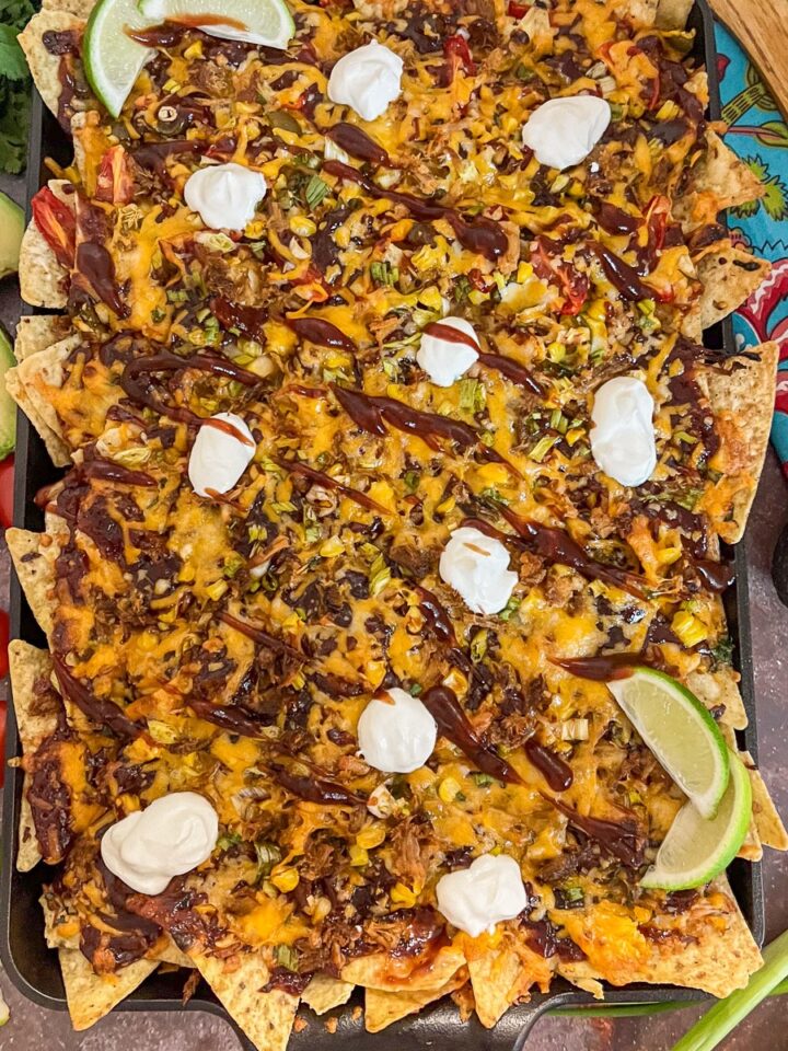 Loaded pulled pork nachos on a cast iron baking sheet with sour cream, barbecue sauce, and fresh toppings