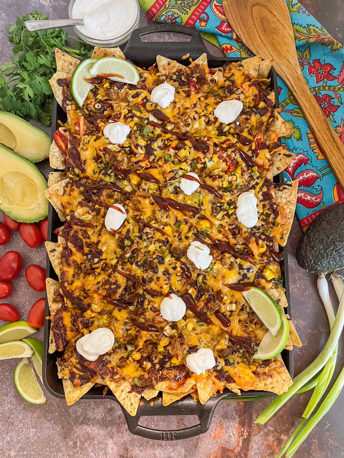 Overhead view of loaded pulled pork nachos on a cast iron baking sheet with sour cream dollops, barbecue sauce drizzle, avocado, and fresh garnishes
