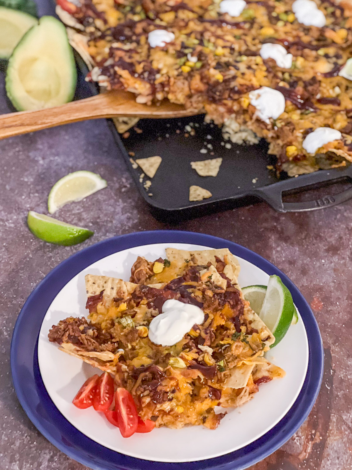 A serving of pulled pork nachos on a blue plate with lime wedges and tomatoes, cast iron baking sheet in background