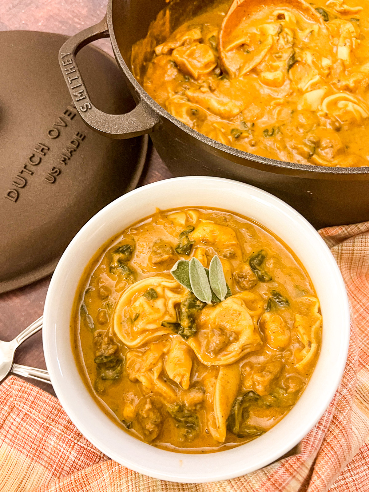 Sausage butternut squash tortellini soup in a white bowl with sage leaves garnish next to a Staub dutch oven