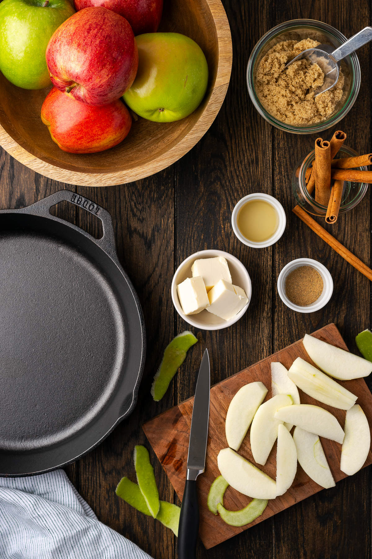 cast iron cinnamon apples ingredients on a dark wood surface with apples brown sugar butter cinnamon and a Lodge skillet