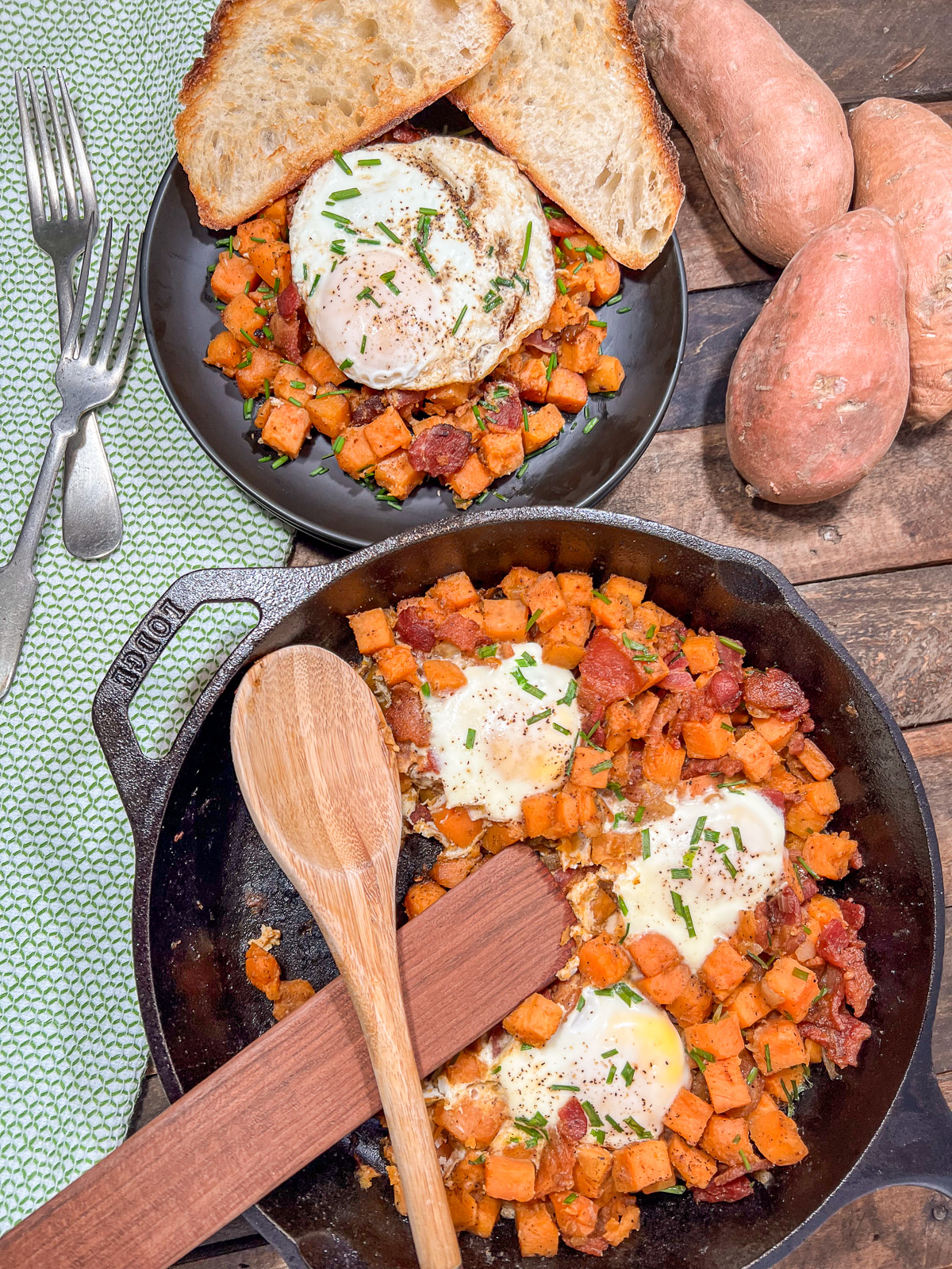Overhead view of sweet potato hash in a Lodge cast iron skillet with a plated serving, toast, and whole sweet potatoes on a rustic wood table.