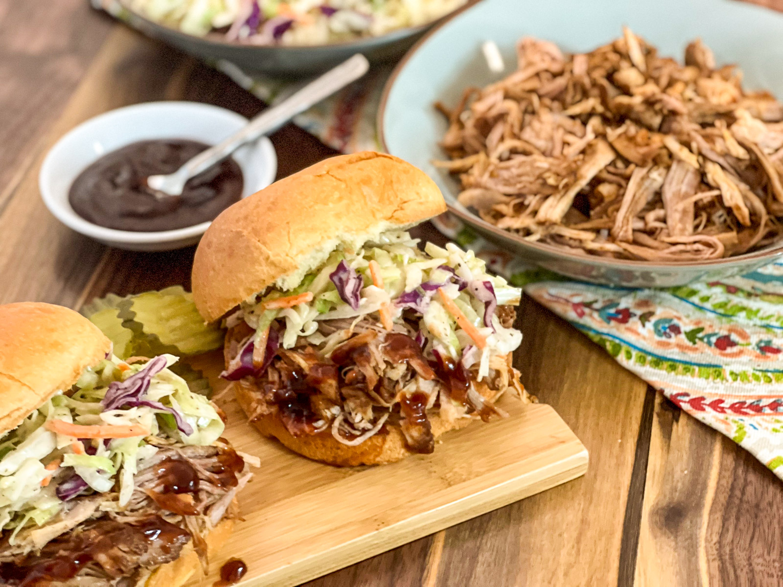 Pulled pork sandwiches with coleslaw on a cutting board with a bowl of shredded smoked pork.