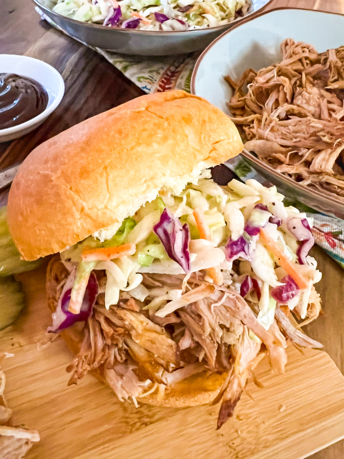 Pulled pork sandwich with coleslaw, BBQ sauce, and bowl of shredded pork on a wood table.