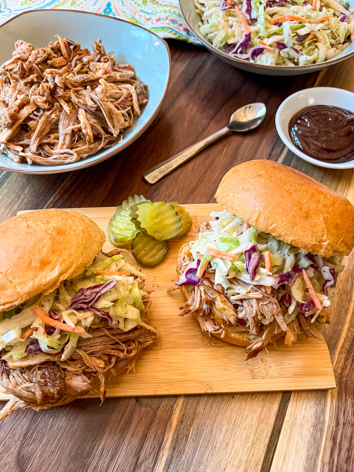 Two pulled pork sandwiches with coleslaw on a cutting board, bowl of shredded pork and BBQ sauce.