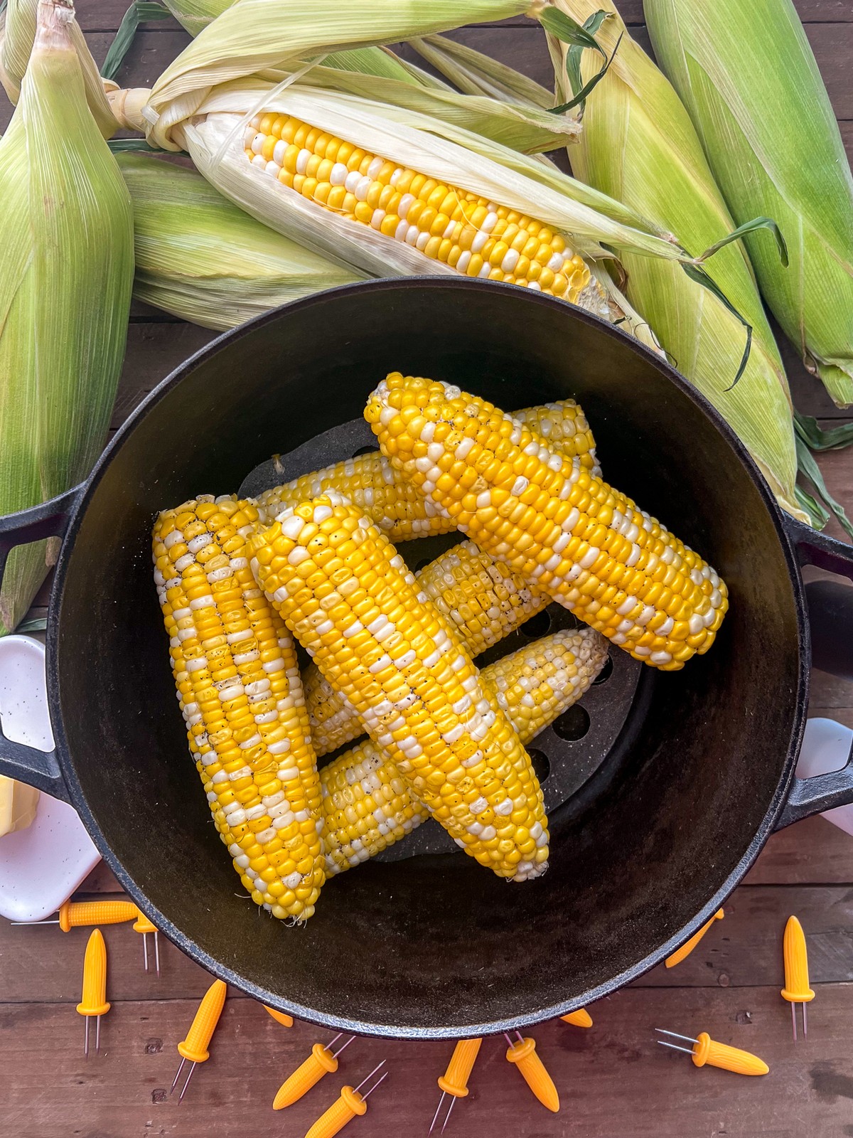 Freshly steamed corn on the cob in a dutch oven with fresh corn in the background along with corn prongs.