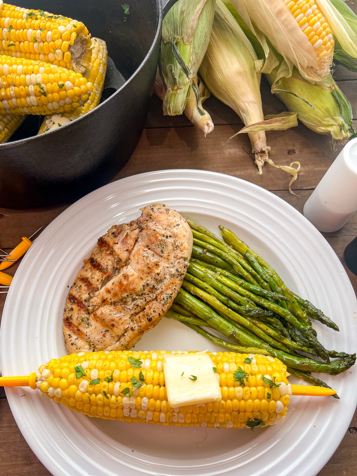Corn on the cob plated with seasoned chicken breast and fresh asparagus on a white plate with more fresh corn, cooked and uncooked in the background.